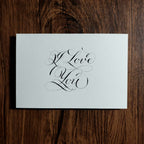 "I Love You" hand-lettered in copperplate calligraphy on a white card on a dark wood table